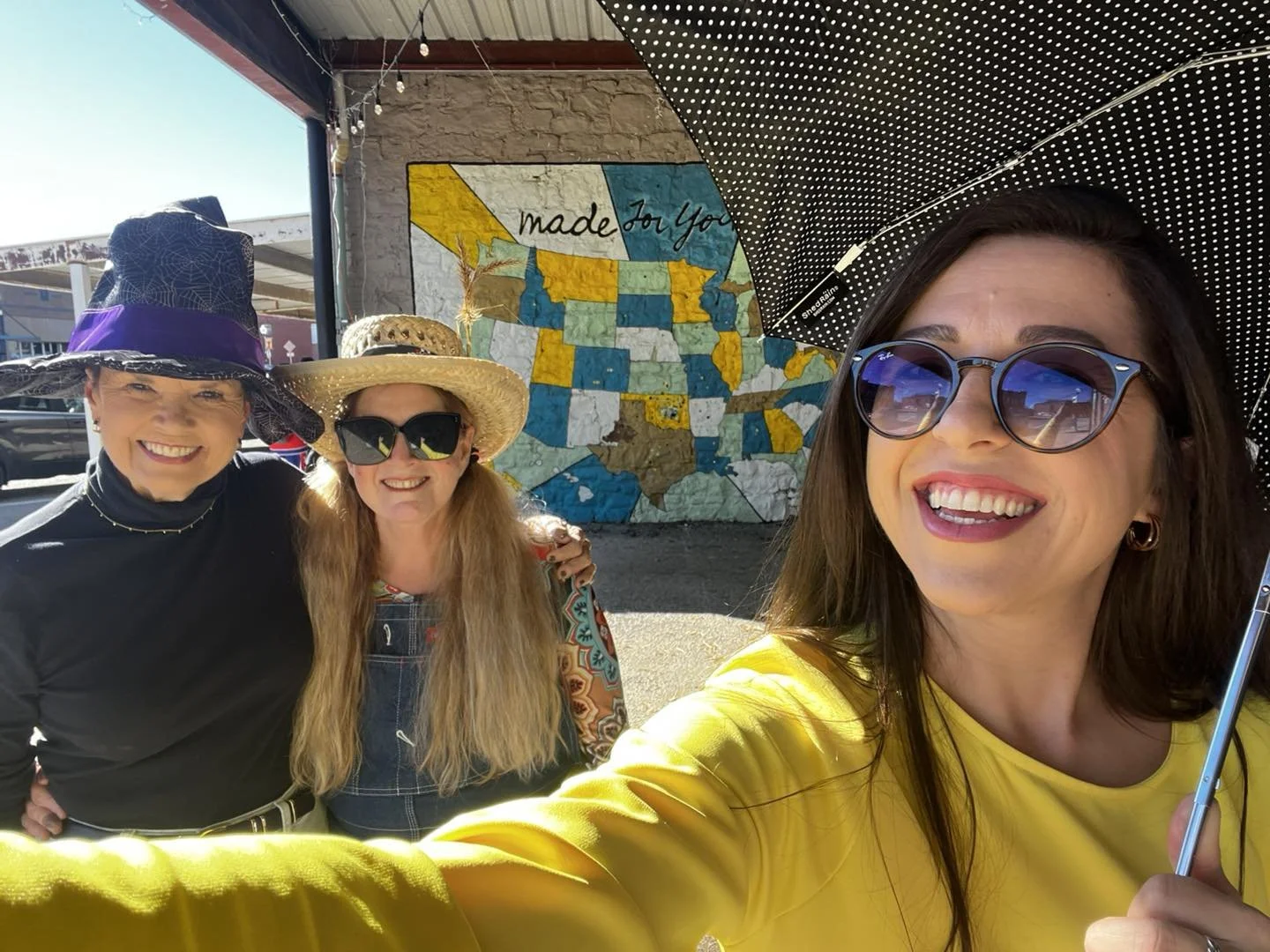 Three women smiling for a selfie outdoors, two wearing large hats and sunglasses, one holding an umbrella. Behind them is a mural of the United States map with the words "made for you".