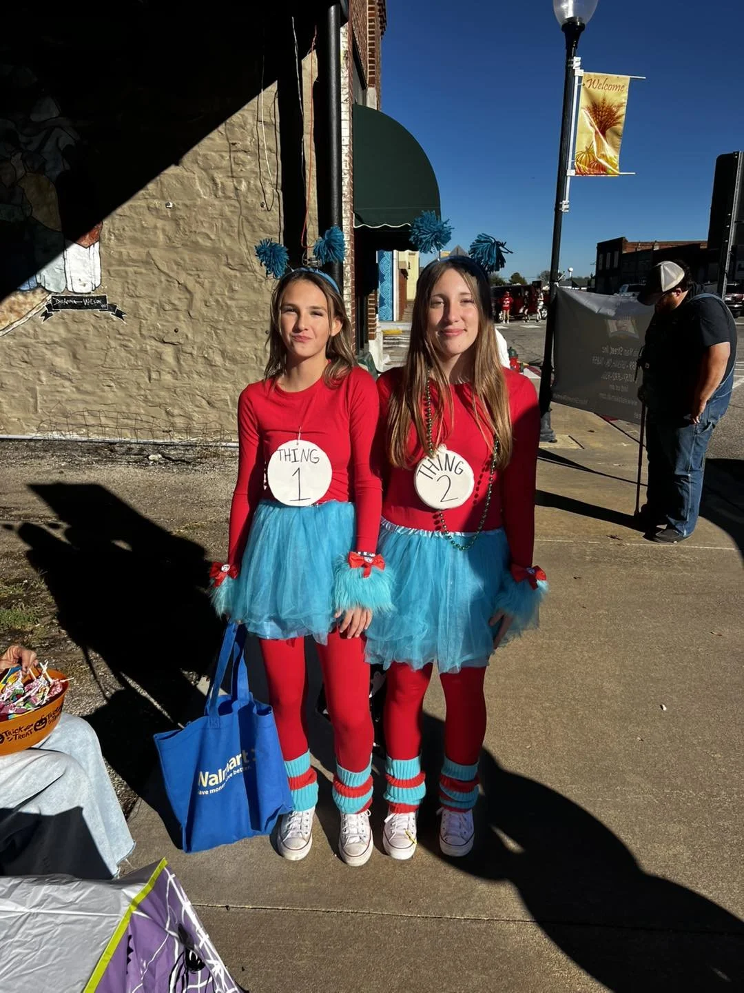 Two young women dressed as Thing 1 and Thing 2 from Dr. Seuss, wearing red tops, blue tutus, red tights, and accessories, standing on a sidewalk in a festive outdoor setting.
