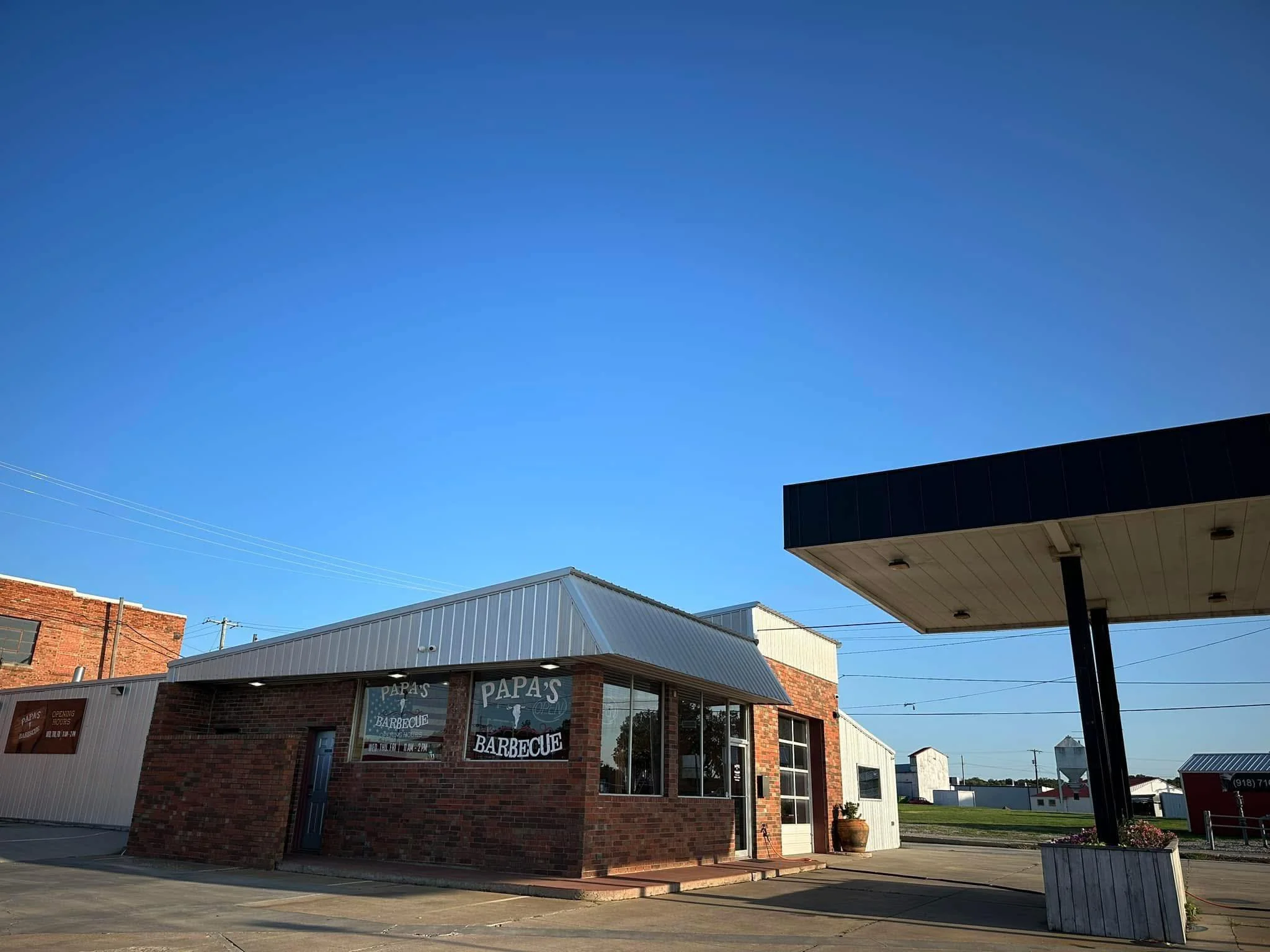 A small brick building with signage that reads 'Papa's Barbecue' and large windows, situated next to a parking lot and a black awning at a gas station, under a clear blue sky.