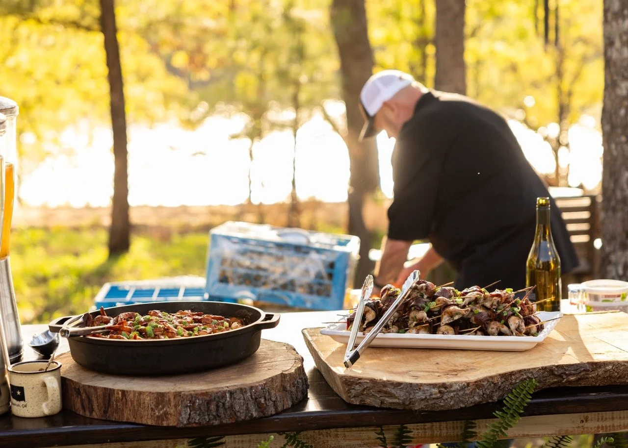 Outdoor food setup with grilled skewers and a dish on a rustic table in a wooded area during daylight.