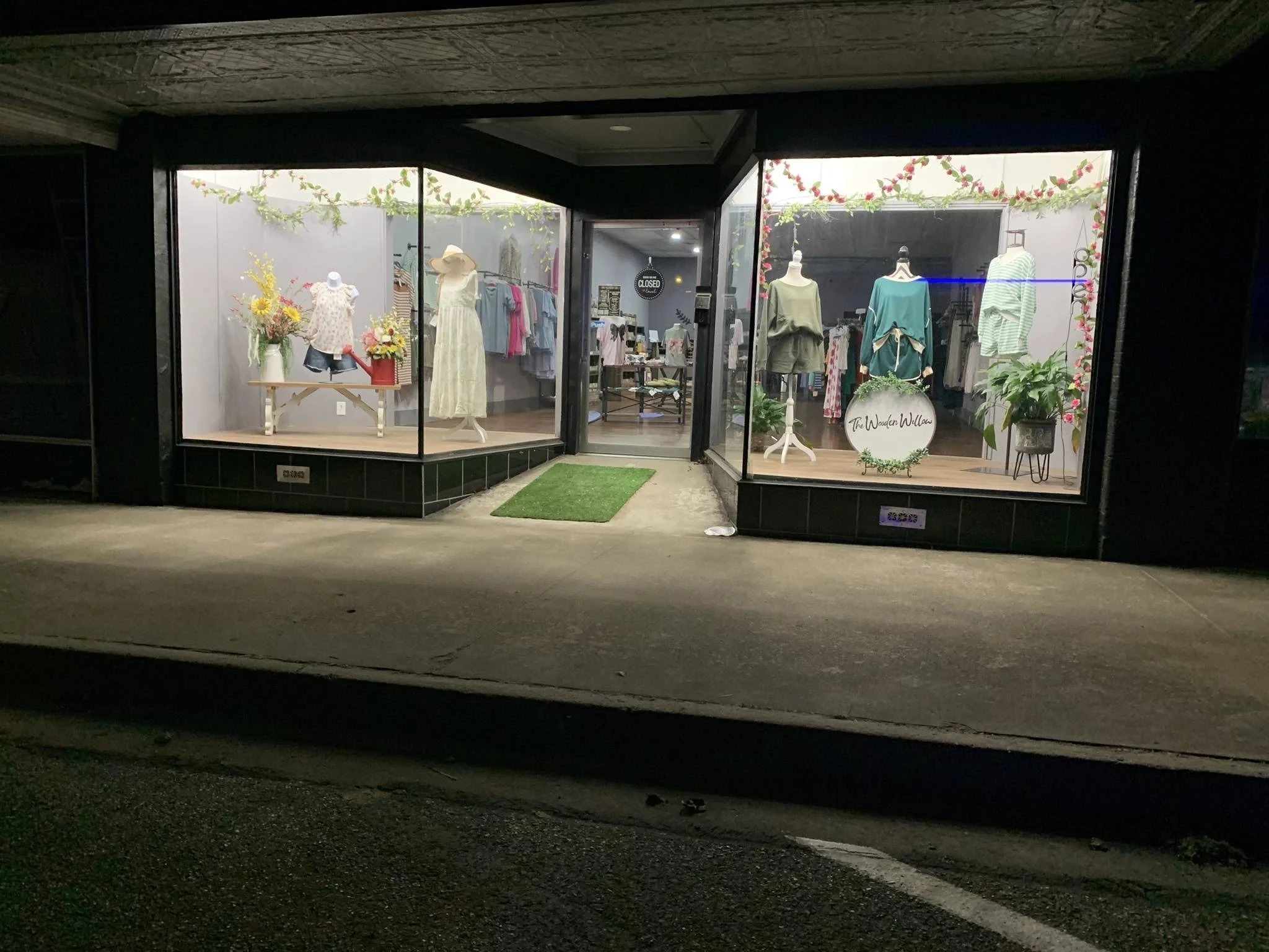 Store display window with mannequins wearing summer clothing, decorated with flowers and vines, night-time view.