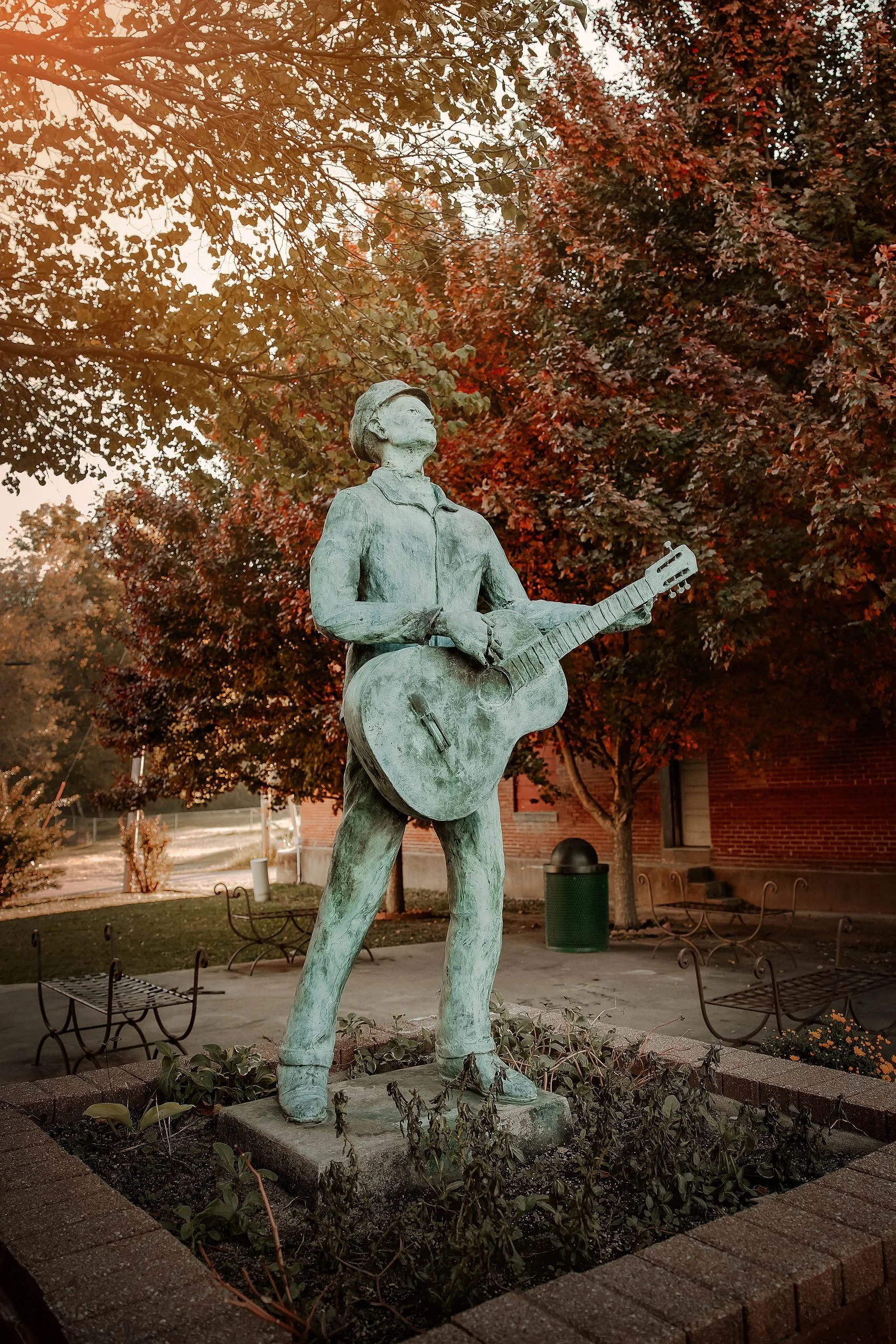 A bronze statue of a woman holding a guitar, standing on a concrete base in an outdoor setting with trees and benches. The woman appears to be looking upward, surrounded by autumn-colored foliage.