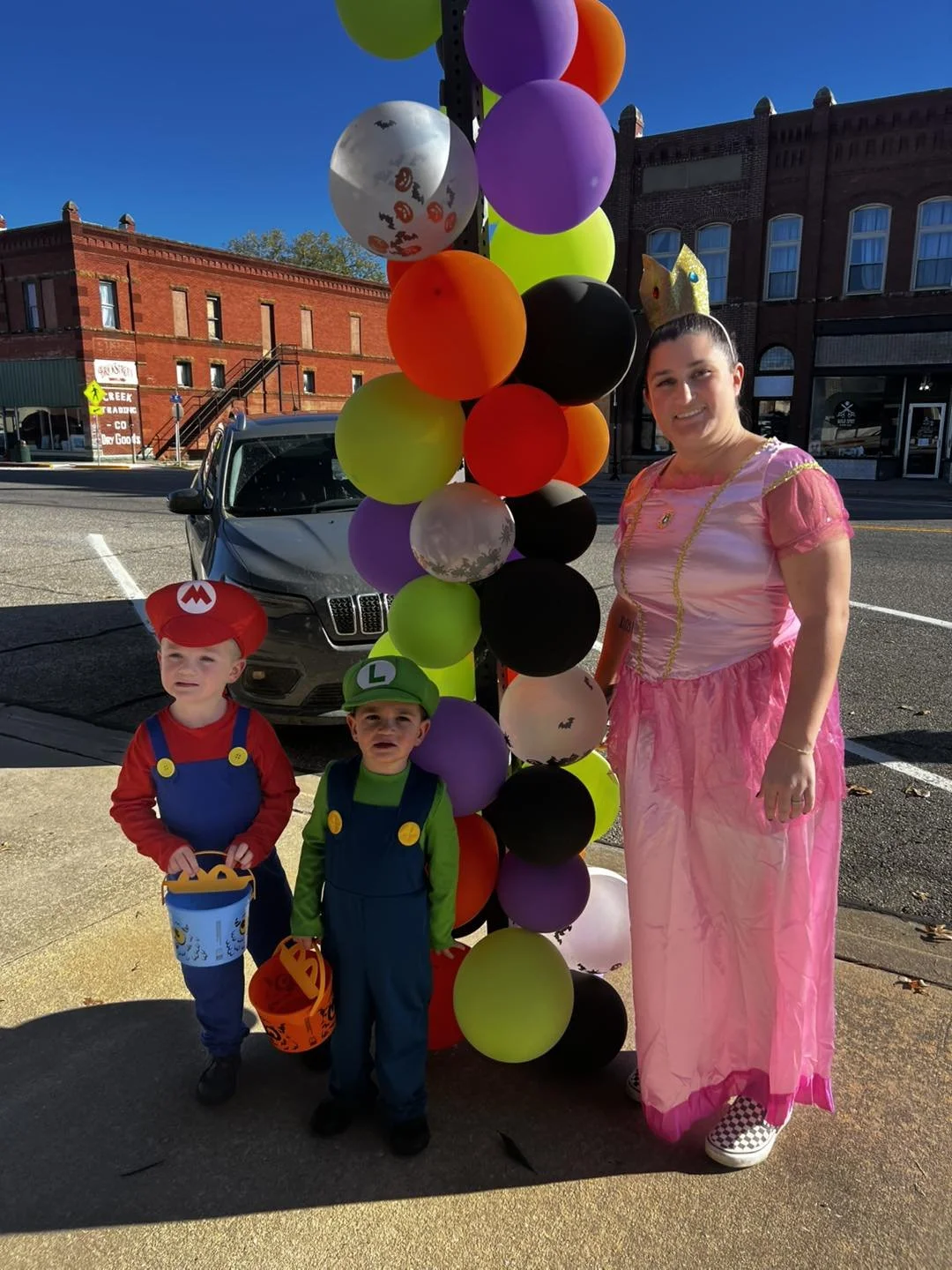 A woman dressed as a princess in pink beside two children dressed as Mario and Luigi holding pumpkin buckets, standing in front of colorful balloons on a city street.