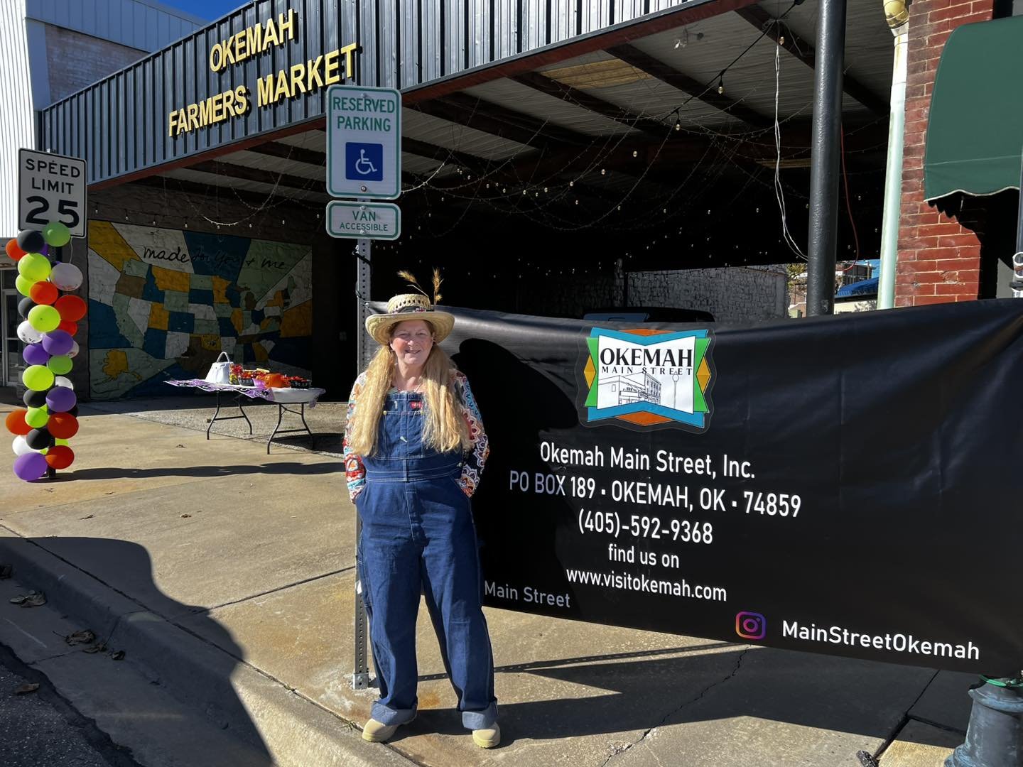 Woman wearing overalls and a straw hat standing next to a sign for Okemah Main Street. The sign includes contact information and social media icons, with a display of balloons on the left and a black banner.