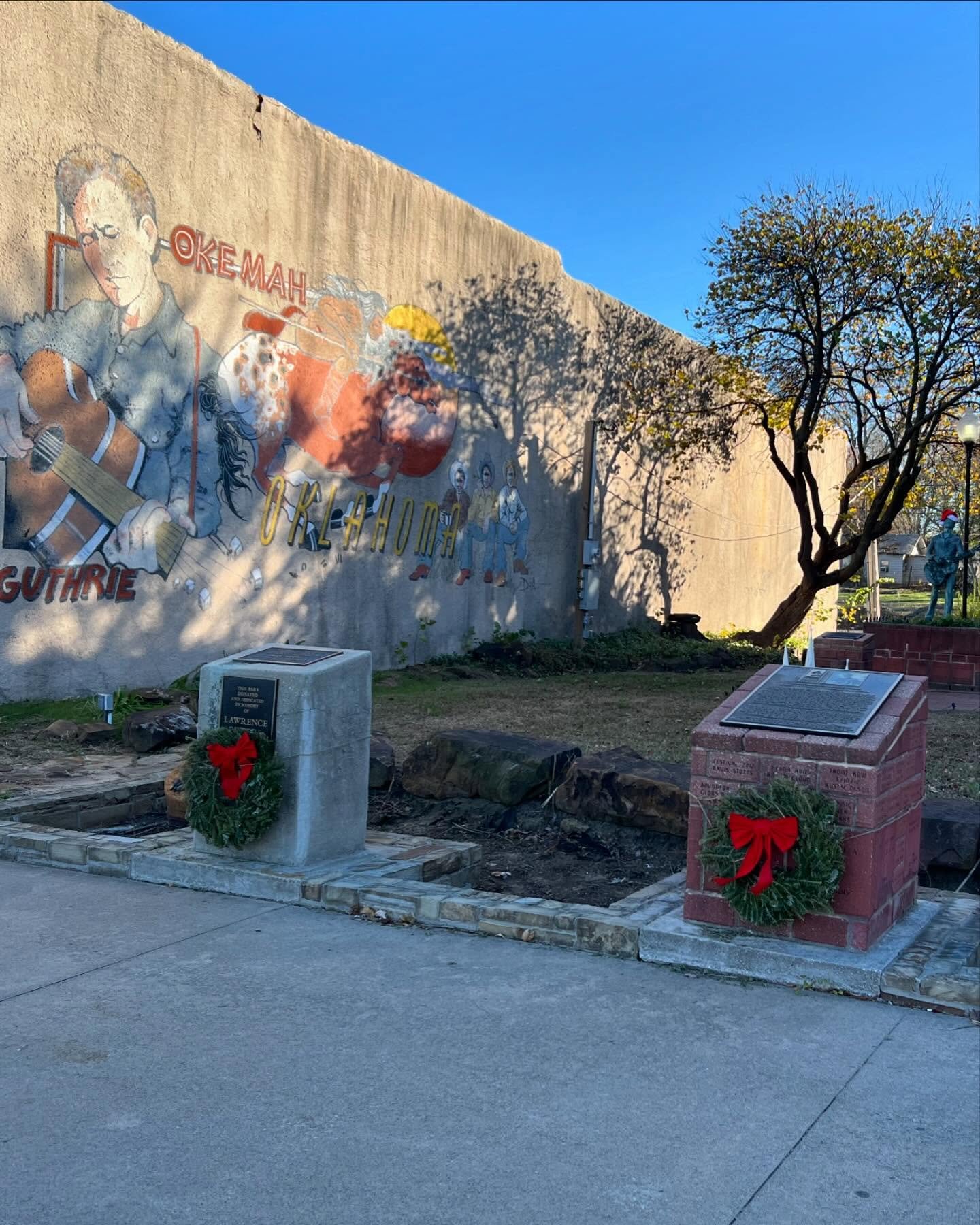A mural on a beige wall depicting a man playing a guitar, a woman holding a guitar, and three children dressed in historical clothing, with the words 'Okemah' and 'Guthrie' visible. In front of the mural are two plaques with holiday wreaths, set on s