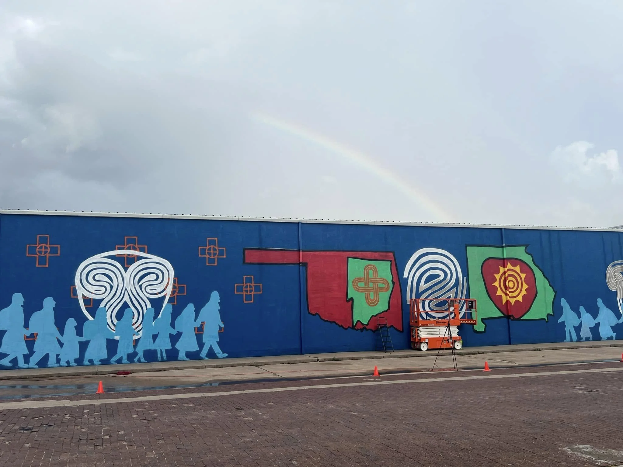 A colorful mural on a blue wall depicting healthcare and state symbols, with silhouettes of people walking, a cherry blossom, a maple leaf with a sun design, and a stethoscope. A rainbow is visible in the cloudy sky above.