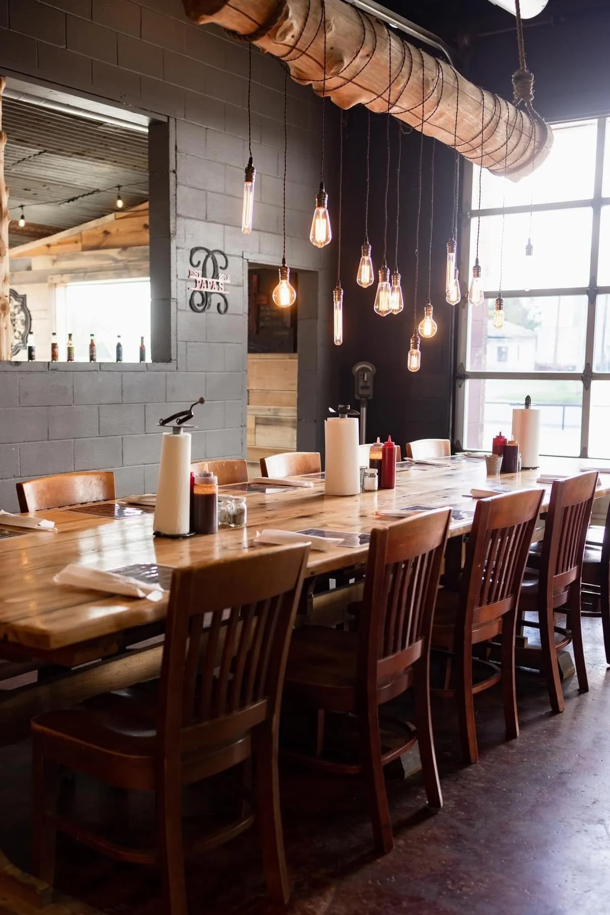 A cozy restaurant interior with a long wooden table set for dining, surrounded by wooden chairs. Overhead, vintage-style Edison light bulbs hang from the ceiling. Large windows on the right let in natural light, and there are condiment bottles on the