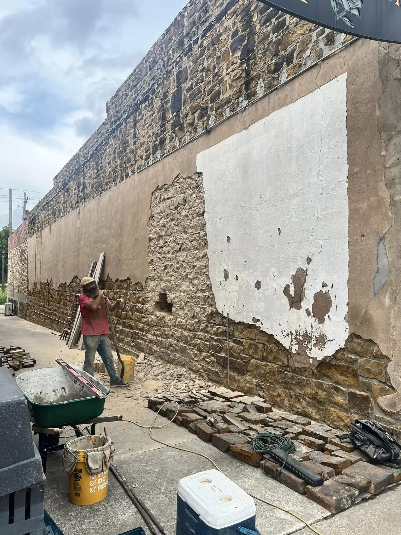 A construction worker with a beard and hat bricklaying on a partially restored stone wall. Various tools and materials are scattered on the ground.