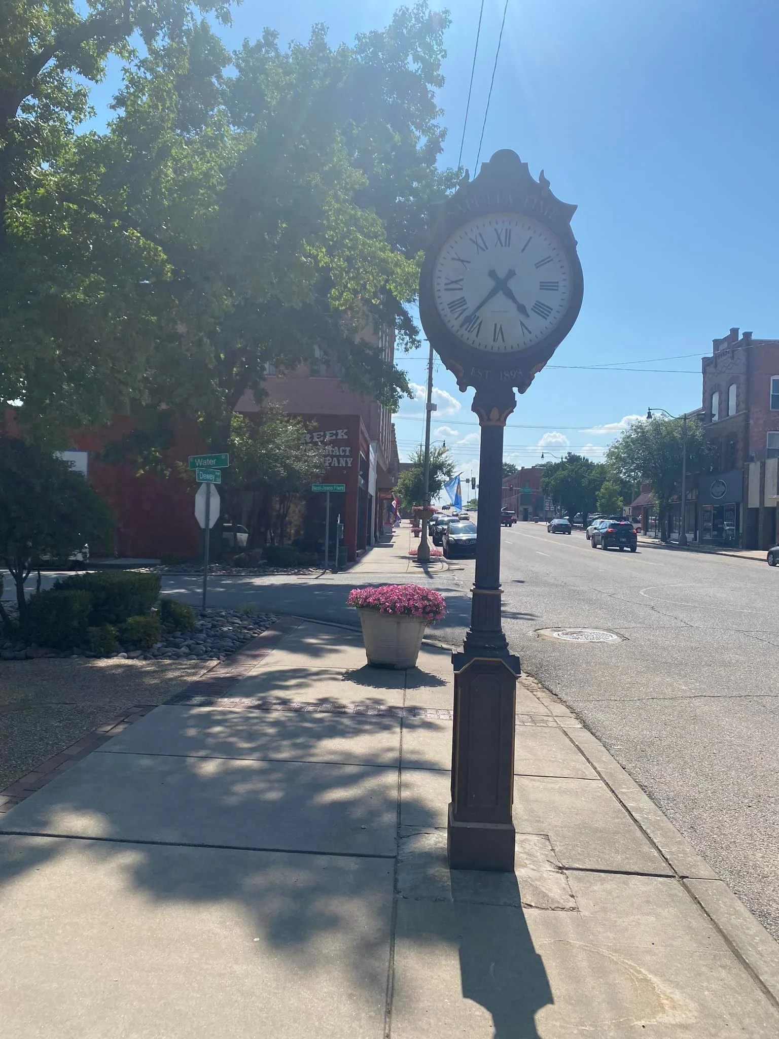 A street view with a vintage-style clock on a tall post, showing approximately 5:42, on a sunny day with clear skies. There are trees, a flowerpot with pink flowers, and several buildings and cars in the background.