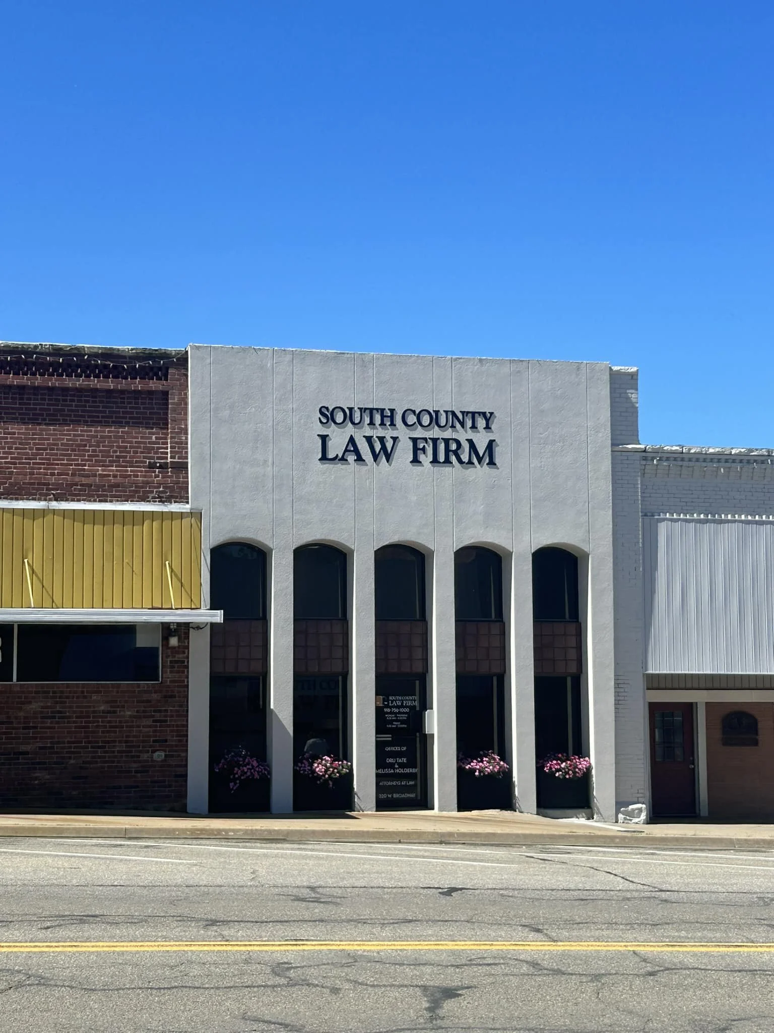 Front facade of a building labeled 'South County Law Firm' with three arched windows, flower planters, and a blue sky overhead.