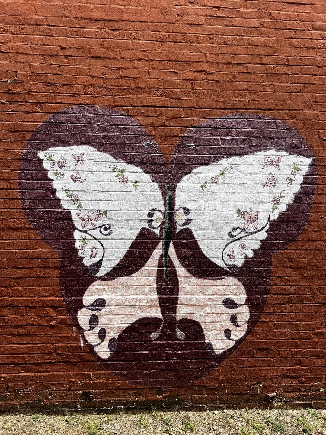 Street art of butterfly-shaped heart painted on a brick wall, with white and brown wings decorated with butterflies and floral patterns.