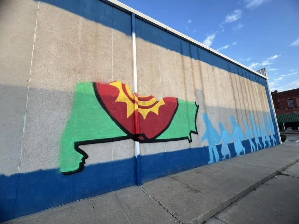 Graffiti mural on a wall depicting a red, yellow, and green corset on a stylized green silhouette, with blue figures walking in a line beside it.