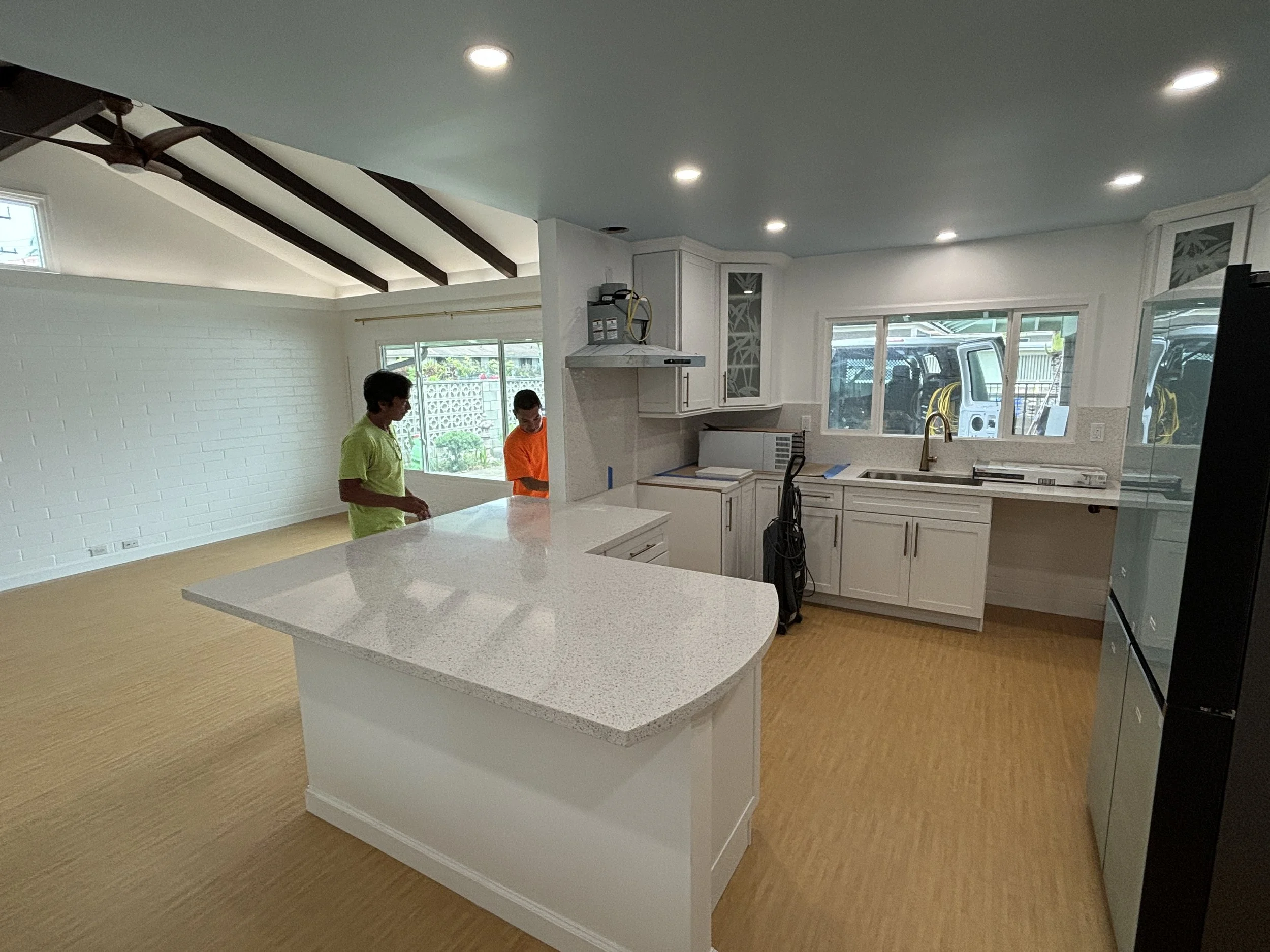Modern kitchen with white cabinets, a large island with a white countertop, a black refrigerator, and a window overlooking a driveway with cars. Two children are standing in the open space.