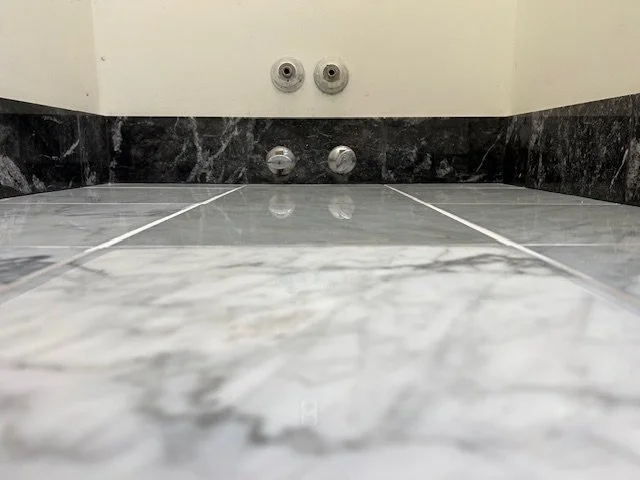 Close-up view of two water supply valves and two drain outlets on a tiled bathroom floor, with black marble wall tiles in the background.