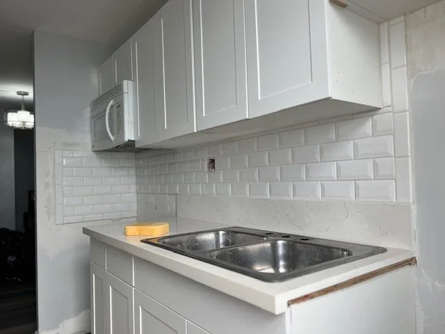 Kitchen countertop with a double stainless steel sink, white subway tile backsplash, white cabinets above, and a microwave installed in the upper cabinets.