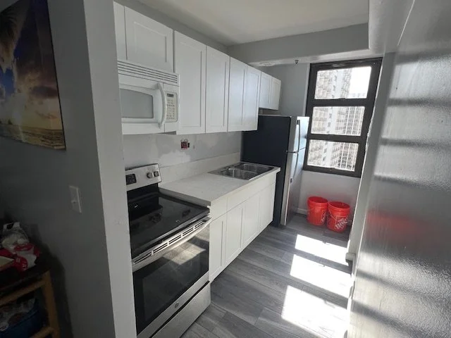 Small modern kitchen with white cabinets, a microwave, stove, refrigerator, and window with city view, two red buckets on the floor.