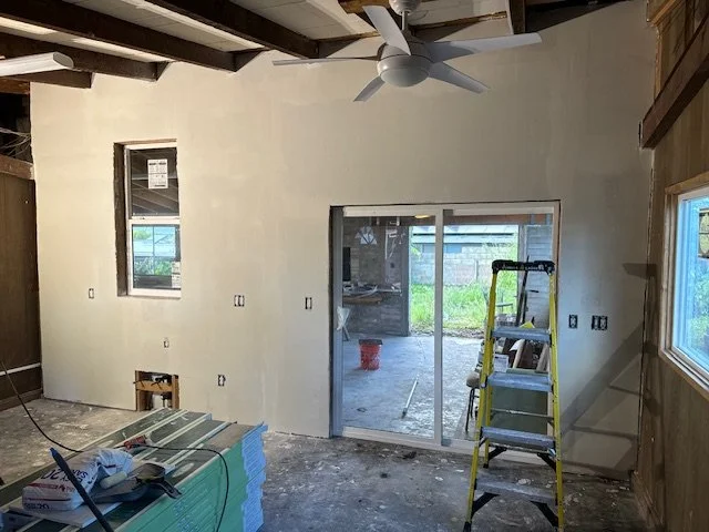 Interior room under construction with unfinished walls, a ceiling fan, a sliding glass door leading to an outdoor area, a window, a yellow step ladder, and construction materials on the floor.