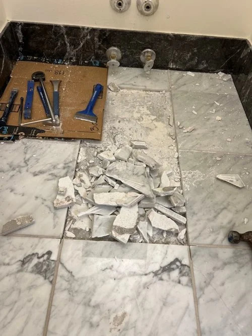 Floor with broken white tile pieces, tools on a cardboard, and pipes against a marble wall.