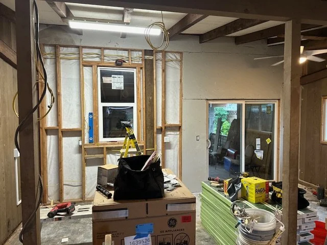 Interior of a room under construction with exposed wall framing, a partially installed window, and construction tools and materials, including a ladder, a black bag, stacked green insulation panels, and boxes.
