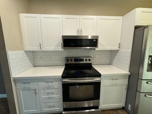 Modern kitchen with white cabinets, stainless steel microwave, and oven, white countertop, white subway tile backsplash, and a stainless steel refrigerator.