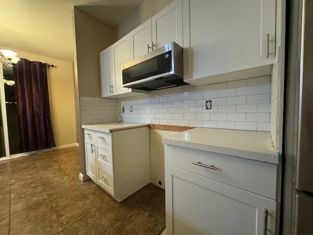 Kitchen with white cabinets, a countertop, a white subway tile backsplash, a black microwave mounted above the stove area, and a sliding glass door with dark curtains in the background.