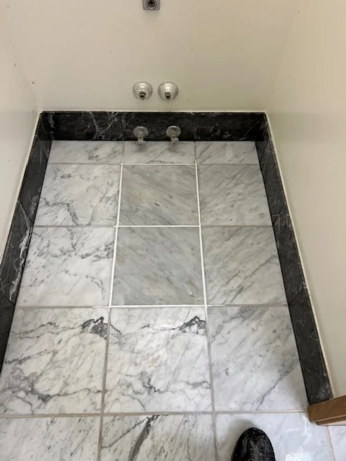 Empty space with tiled marble floor, plumbing fixtures, and a wall with water hookups, likely in a laundry or utility room.