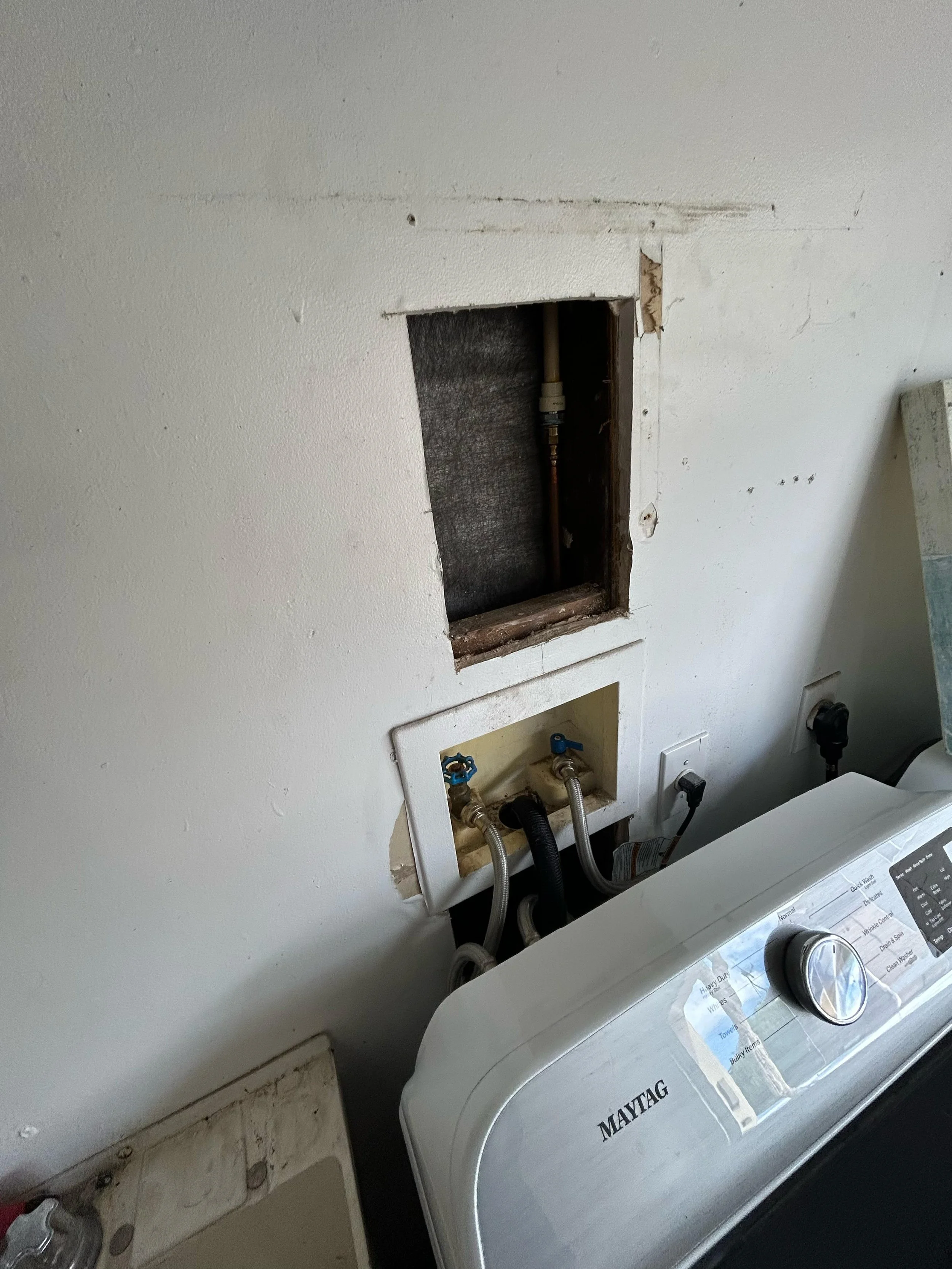 Before drywall repair in a laundry room showing an open wall access behind the washer with exposed plumbing and unfinished drywall that needed patching and restoration.