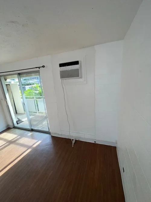 Empty room with hardwood flooring, sliding glass door leading to a balcony, and a wall-mounted air conditioner with a cord hanging down.