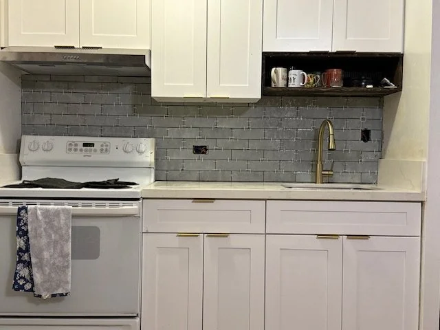 Kitchen with white cabinets, a gray tile backsplash, and a white stove with a towel hanging on the handle. There are cups on a dark open shelf above the sink, which has a gold faucet.