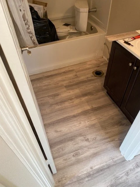 Bathroom with a toilet, black trash bag, and a wooden cabinet on a wood-look vinyl floor. A drain is visible near the cabinet.