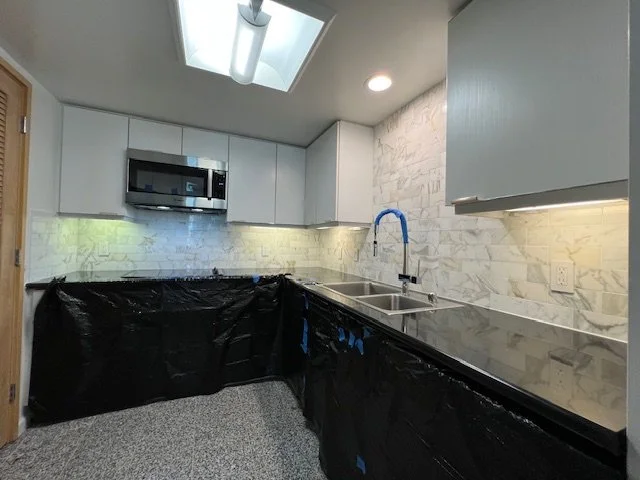Kitchen with white cabinets, marble backsplash, stainless steel microwave, and black plastic covering lower cabinets, with a double sink and a blue faucet.