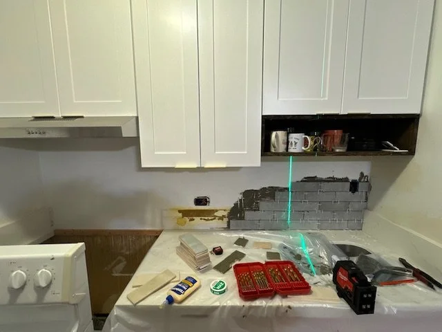 Kitchen countertop with tools and supplies for wall repair, including screws, a level, and a drill, with partially removed backsplash behind white cabinets.