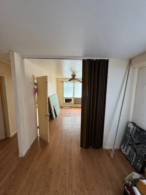 View of an empty room with hardwood floors, a ceiling fan, and a window with blinds. There are some large canvases leaning against the wall on the left, and a black curtain or divider partially separates the space. Unmounted items and a long pole are