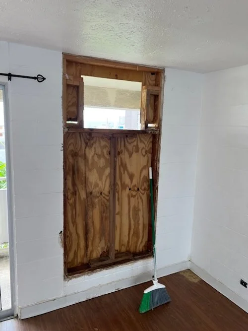 A partially boarded-up window with plywood, next to a broom leaning against the wall, in a room with white walls and wooden flooring.
