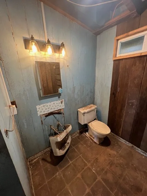 A small bathroom with blue and dark wood paneled walls, a mirror, three wall-mounted light fixtures, a sink, and a toilet. The bathroom appears somewhat outdated and unclean.