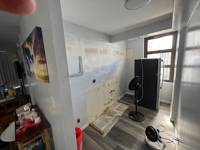 Empty room with a refrigerator, standing fan, window, and a wall with partial paint or wallpaper removal.