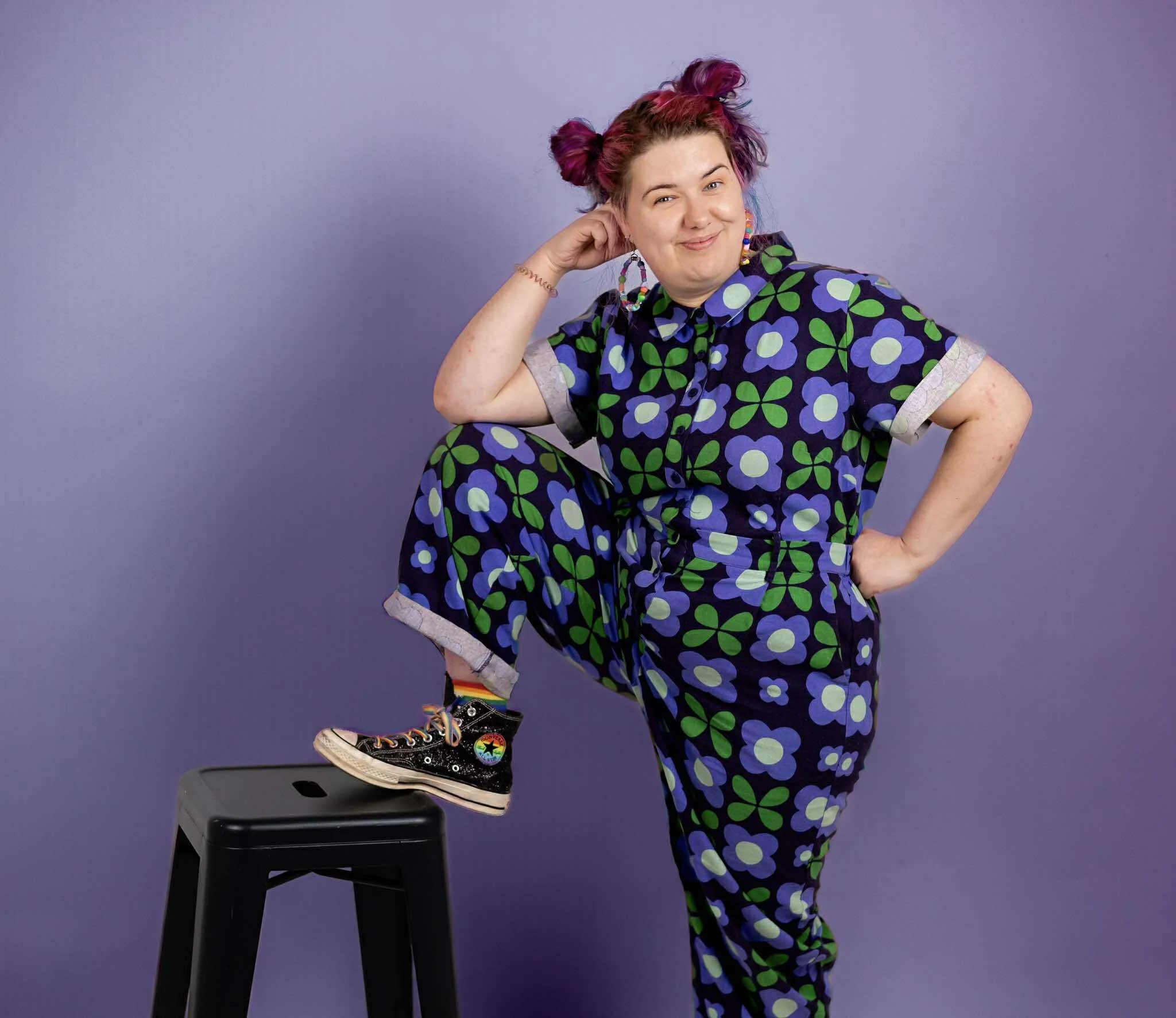 A woman with pink hair styled in two buns, wearing a colorful floral jumpsuit, standing against a purple background. She has one hand on her hip and the other resting on her head, smiling at the camera. Her left foot is raised and resting on a black stool.