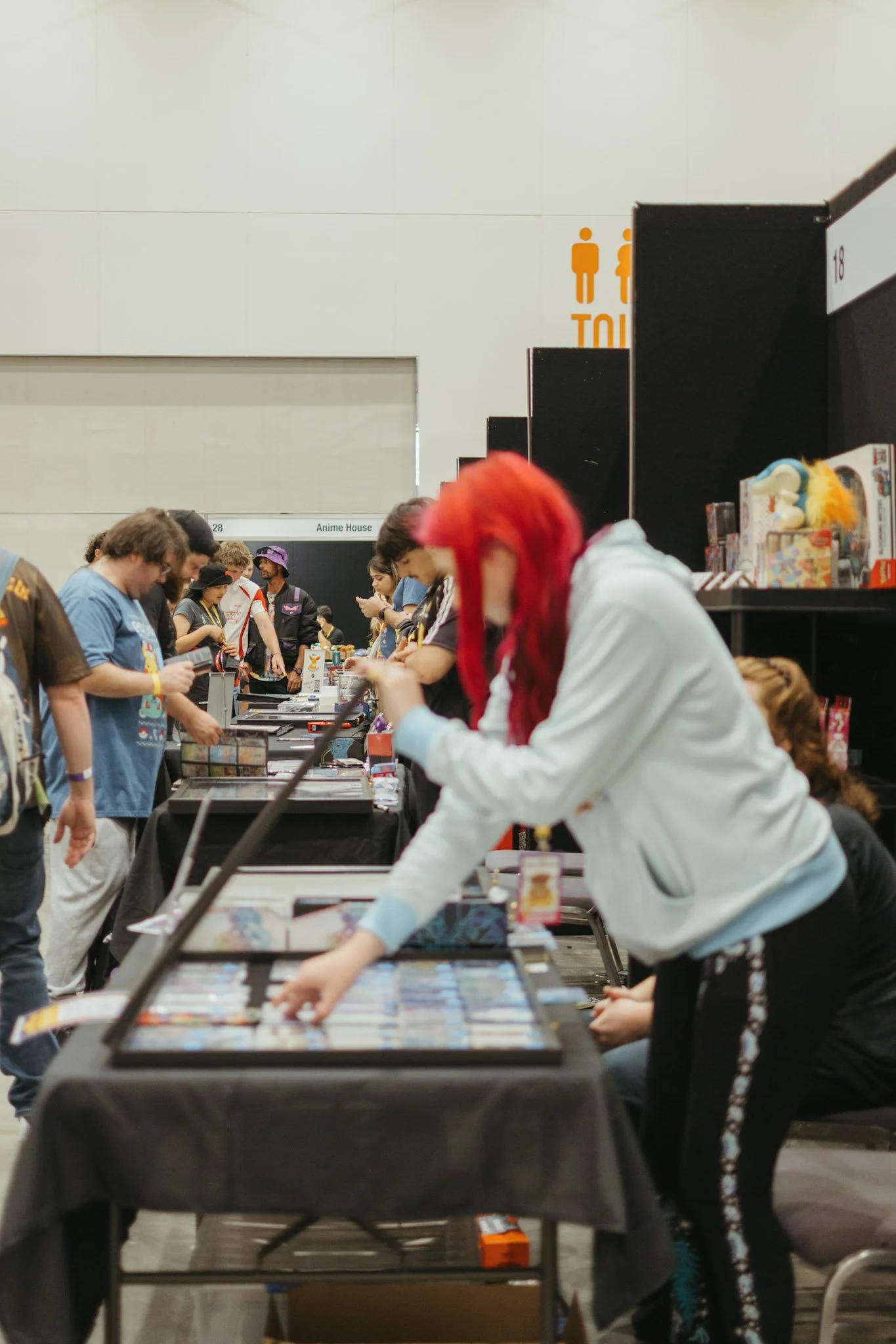 People browsing and purchasing items at a convention or expo booth with a sign indicating restrooms in the background.