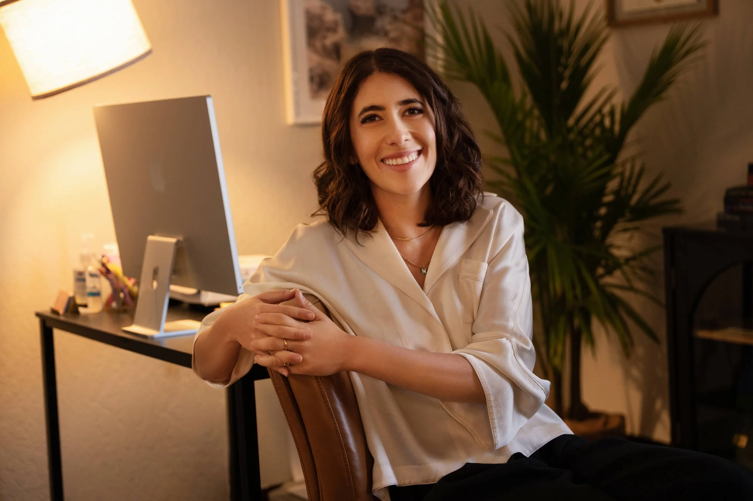 Deb Altman, therapist in Reno Nevada, sits smiling in a cozy therapy office with warm lighting, a large plant in the background, and a desk with a computer and various items.