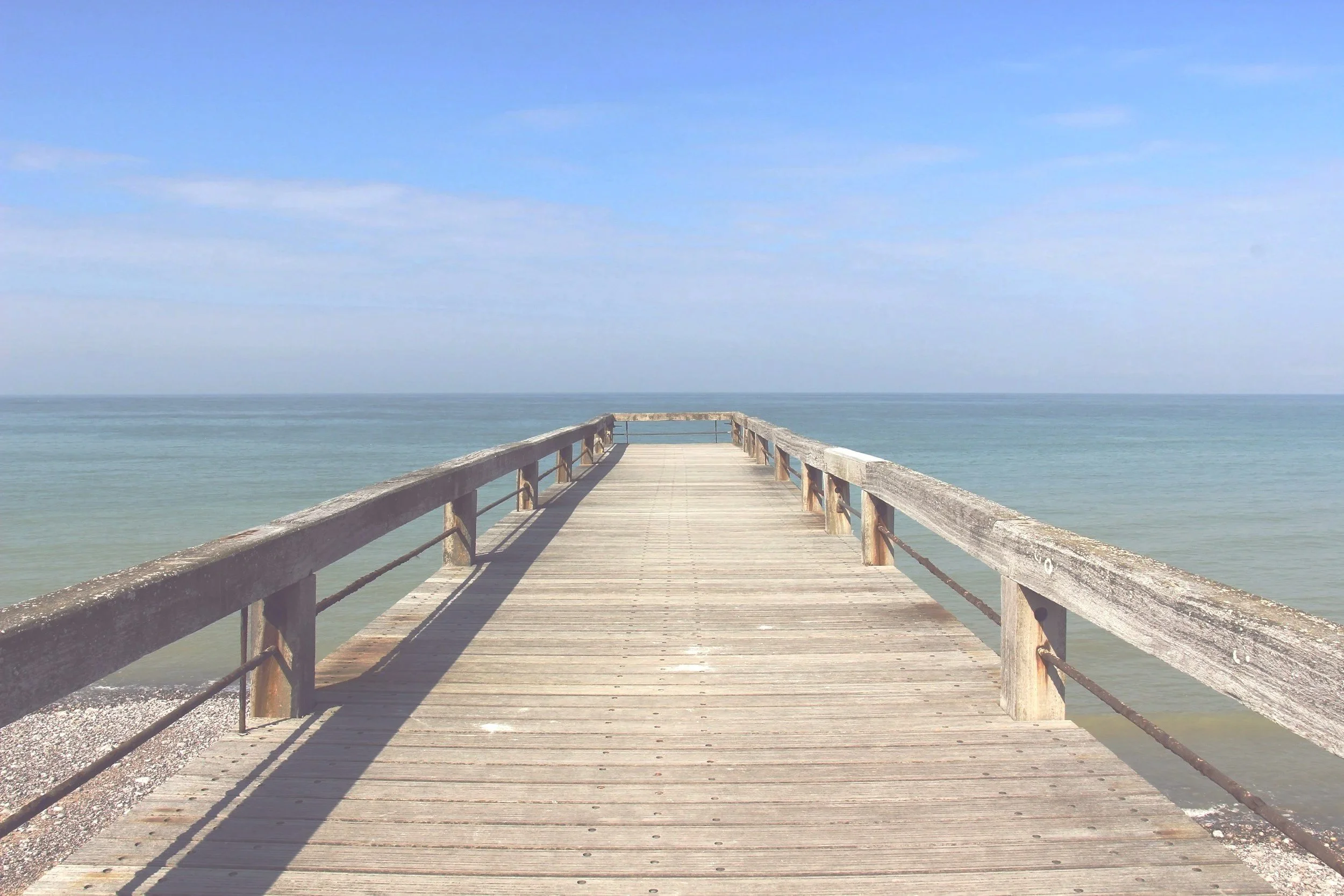A wooden pier extending into the ocean on a clear, sunny day with a mostly blue sky and a few clouds.