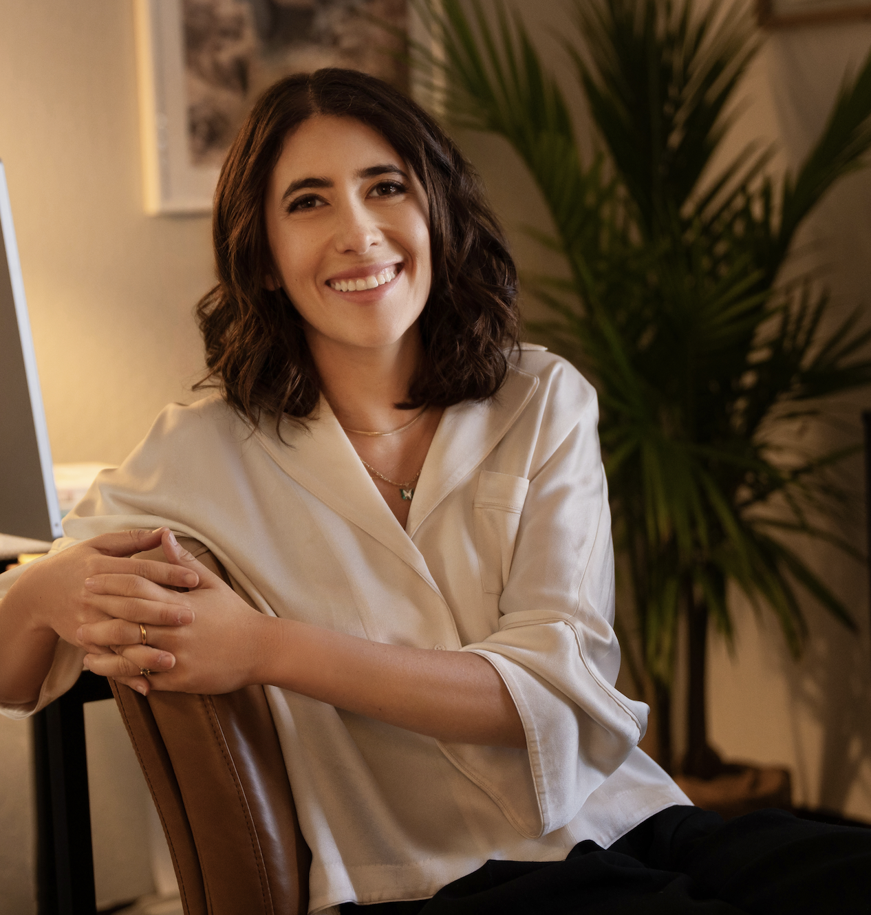 Deb Altman, sitting on a chair, wearing a cream-colored satin shirt, with a large green plant and a wall with a framed picture in the background.
