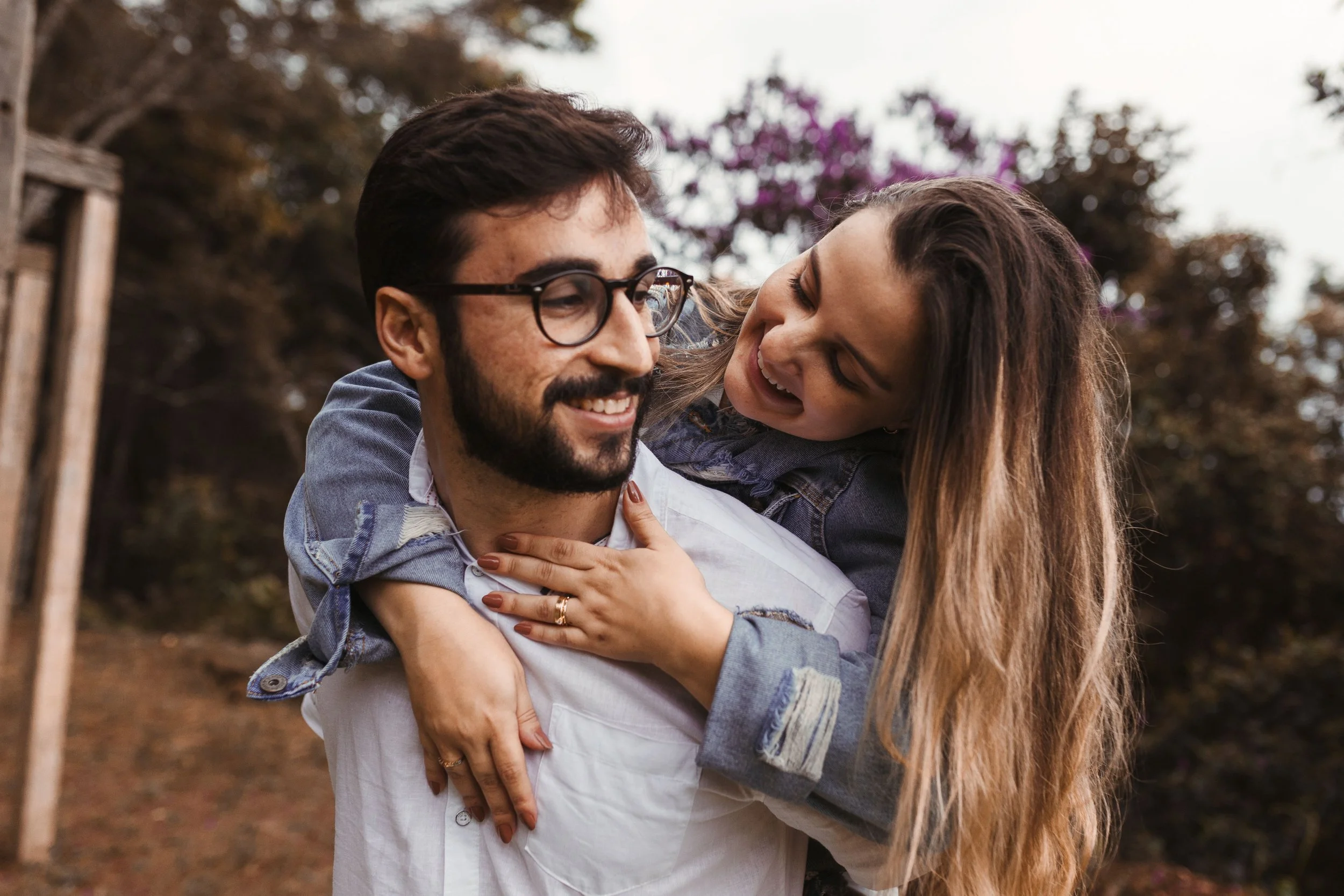 A man with glasses carrying a woman on his back in an outdoor setting with trees and purple flowers in the background.