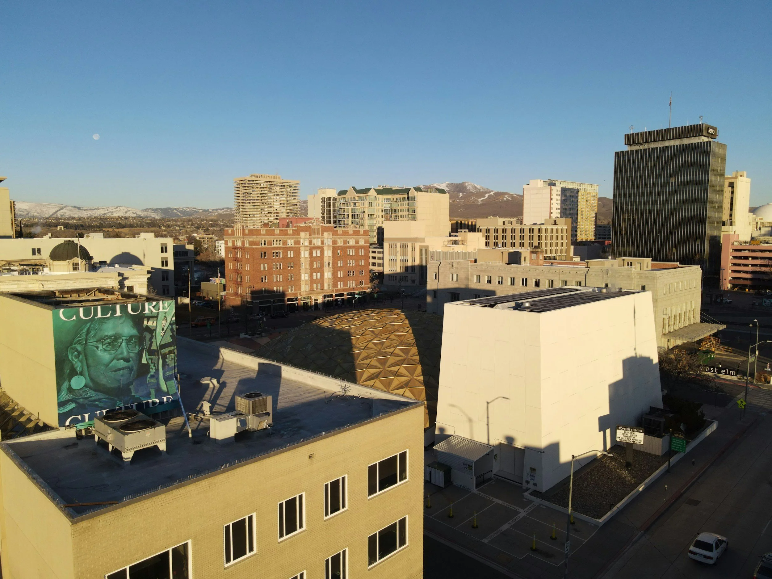 City skyline with various buildings, including a tall black office building, a white modern structure, and a mural on a building's rooftop, with mountains in the background and a clear blue sky.