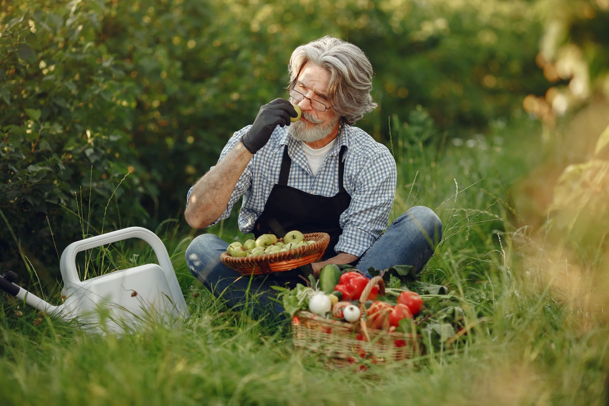 An older man with gray hair and a beard, wearing glasses, a checkered shirt, black apron, and gloves, is sitting outdoors on the grass.