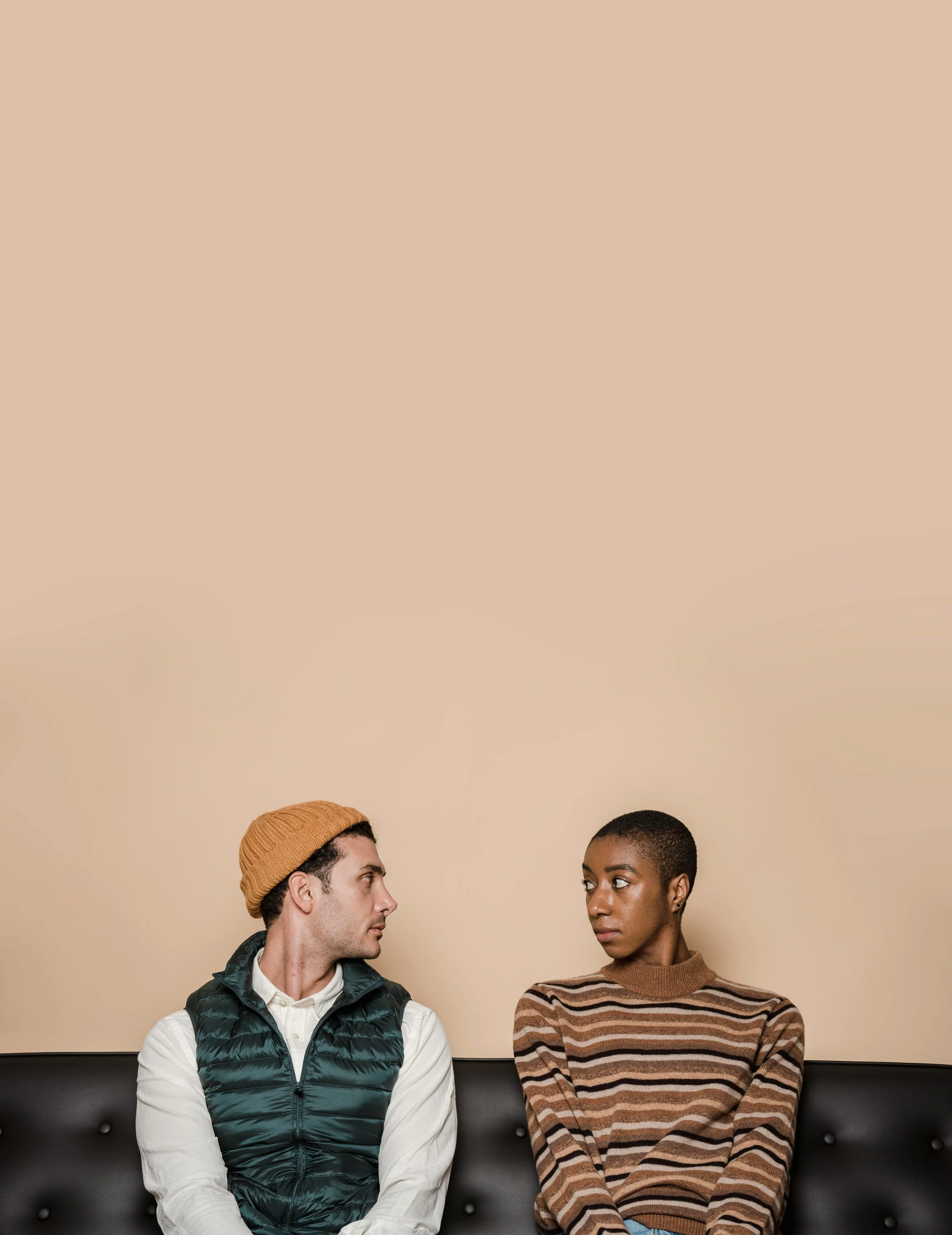A man and woman sitting on a bench against a beige wall, looking at each other. The man is wearing a tan beanie, white shirt, and navy puffer vest. The woman is wearing a striped brown sweater.