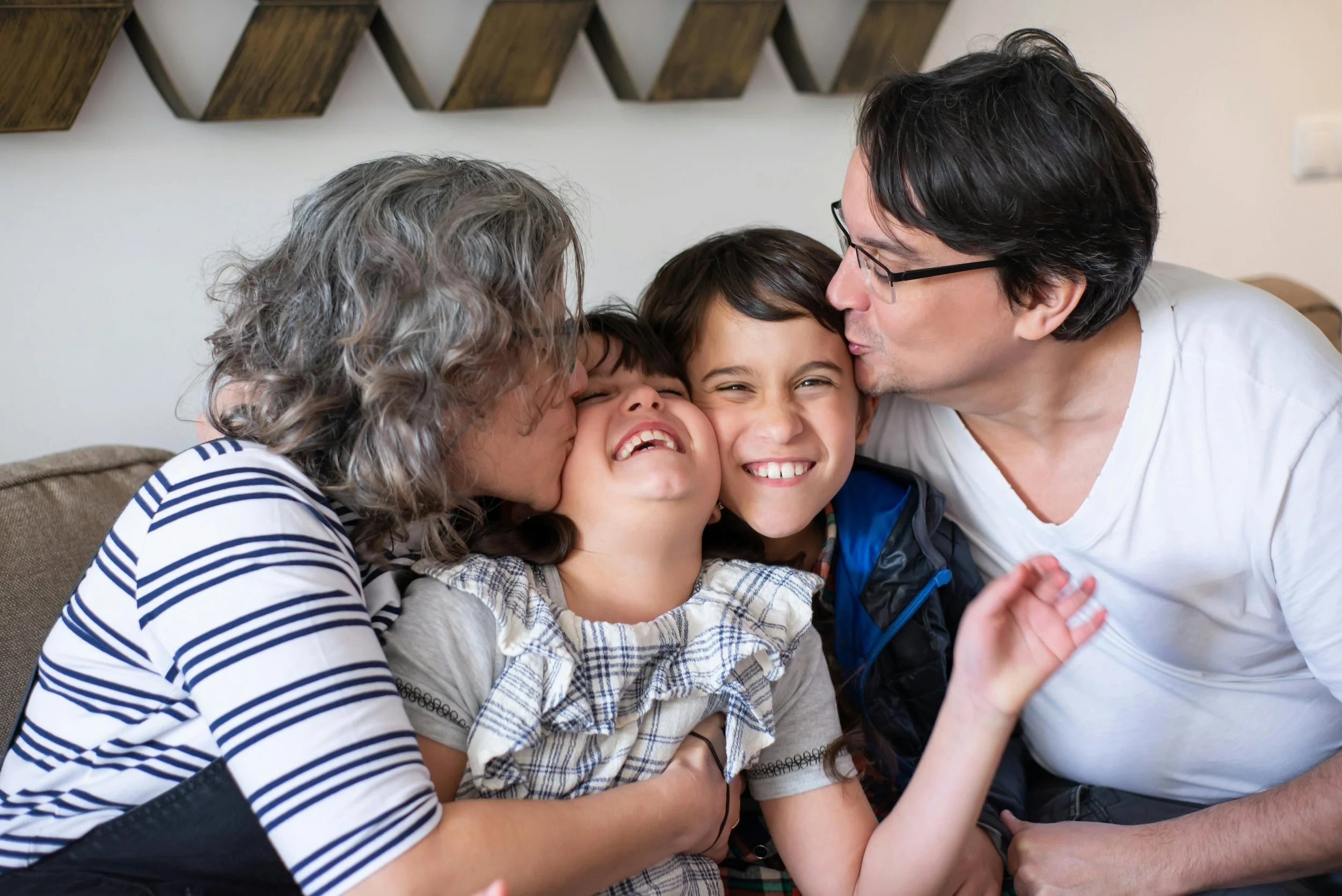 Family of four hugging and kissing each other on a beige sofa in a living room, smiling and enjoying a happy moment.