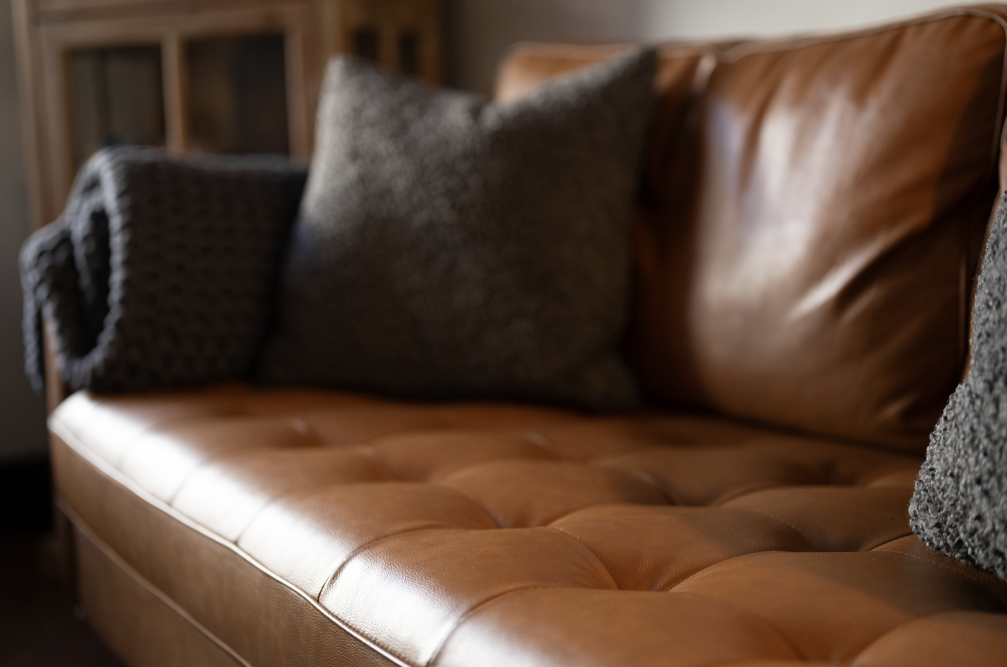 A tan leather sofa with tufted cushions and two gray textured throw pillows, situated in a room with wooden shelves in the background.