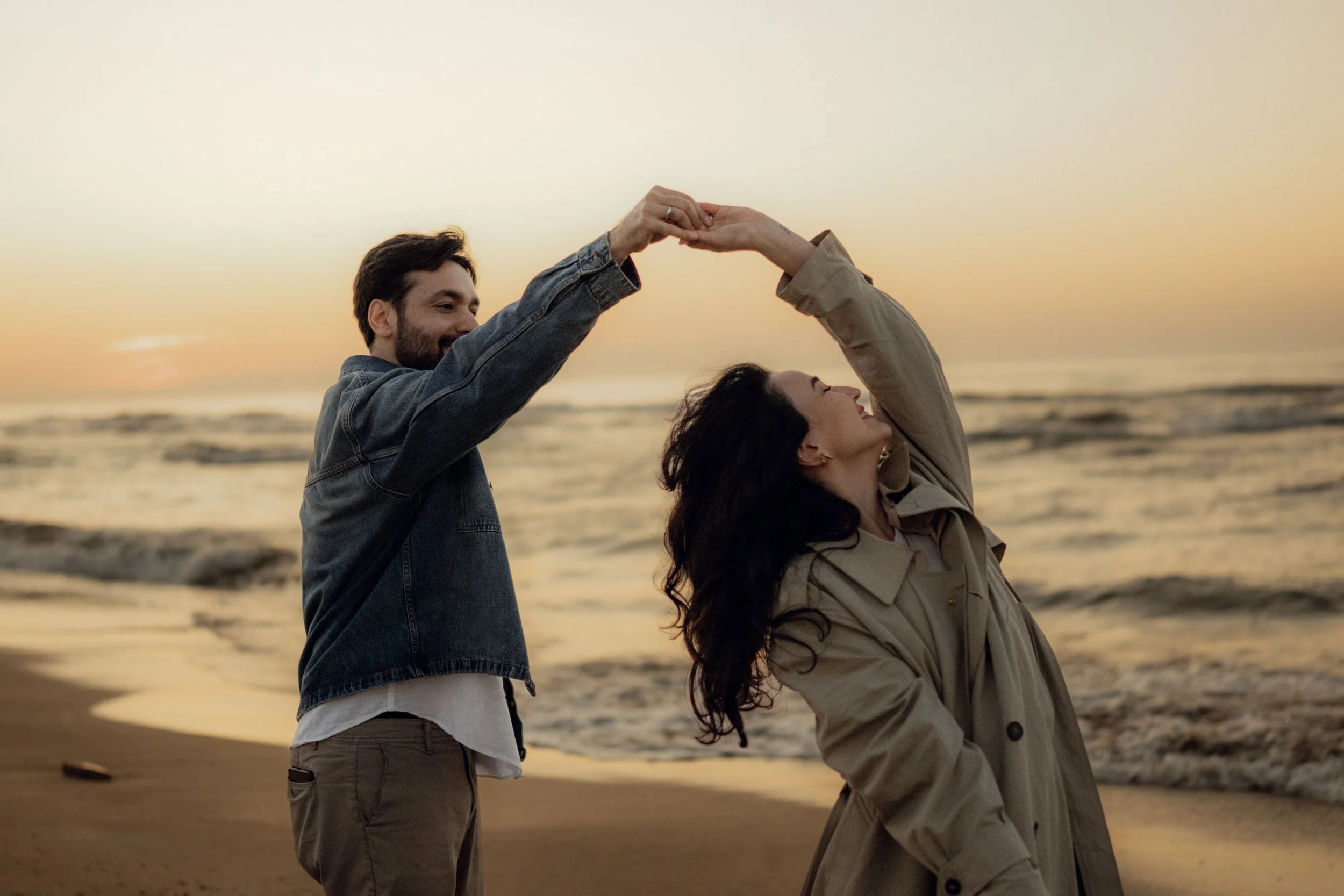 A couple at the beach during sunset dancing and holding hands, with the ocean and sky in the background.