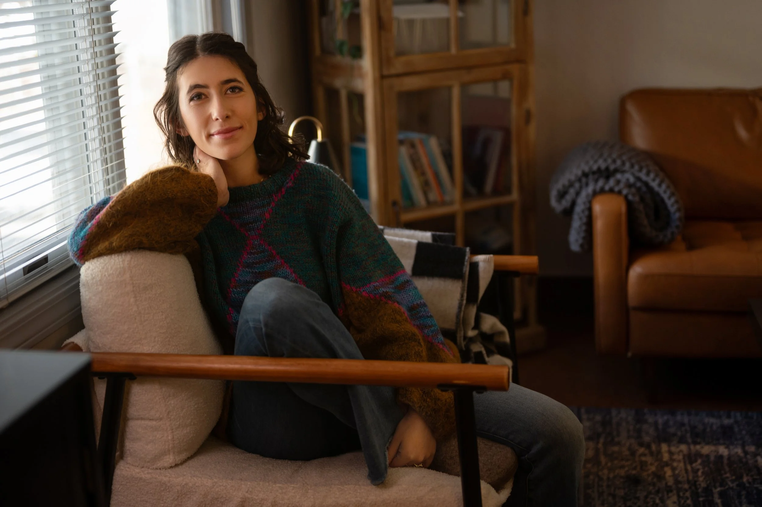Deb Altman, marriage and family therapist in Reno, sitting on a beige armchair near a window with white blinds, inside a cozy therapy office, smiling softly.