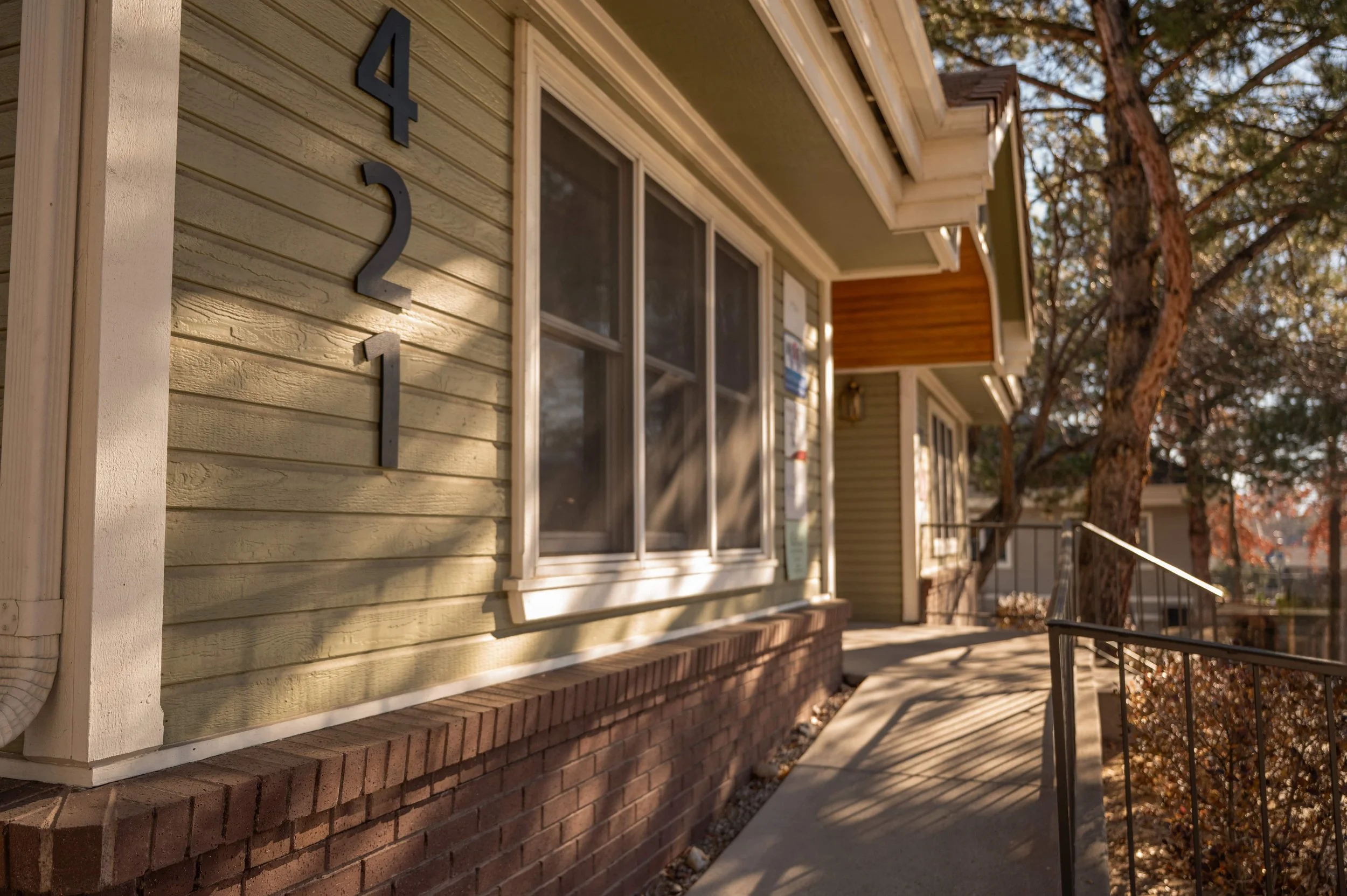Residential building with house number 421, beige siding, decorative brick foundation, and a sidewalk with a metal railing in a suburban neighborhood with trees.